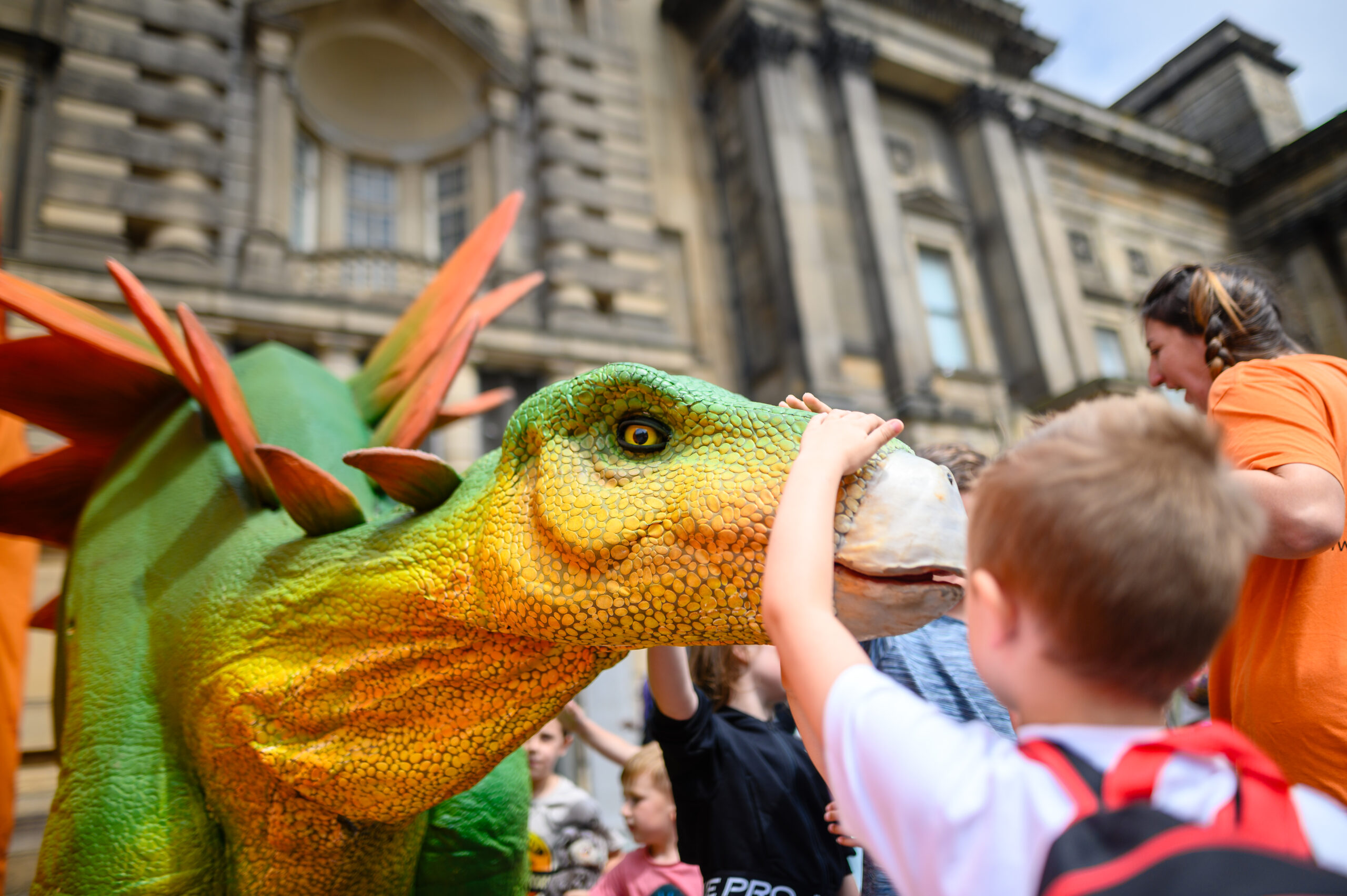 Children interacting with a dinosaur prop at the Dinomania event at World Museum Liverpool, with a large building in the background.