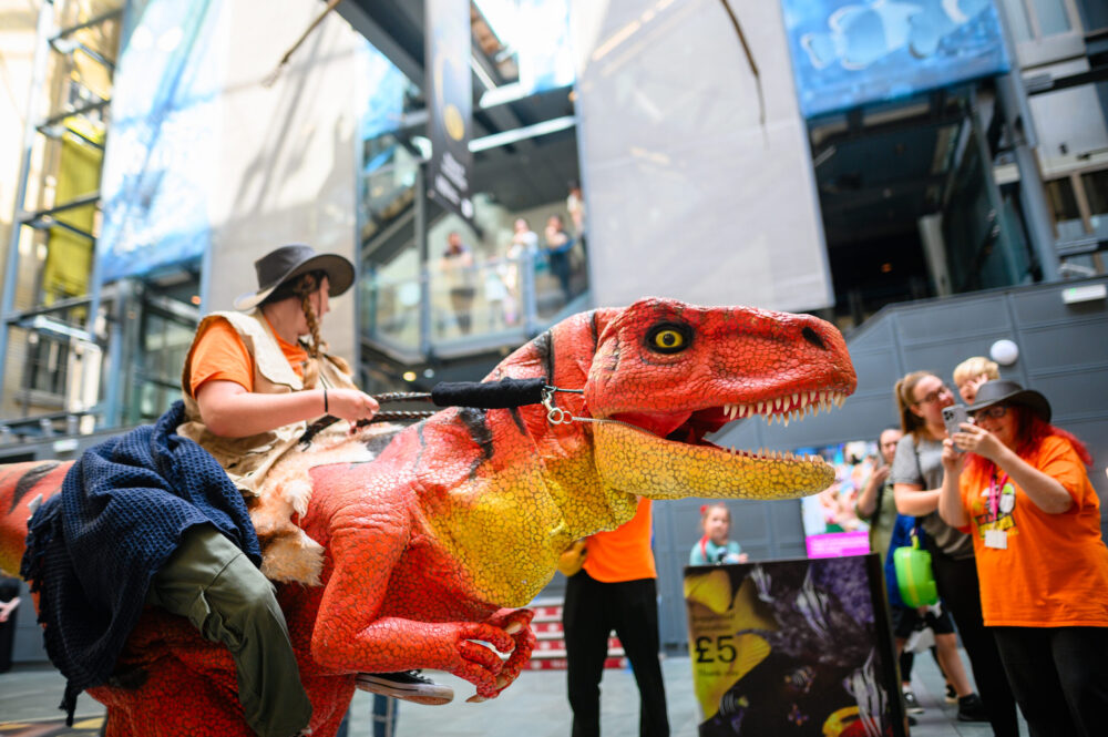 A person dressed as a dinosaur is being ridden by another person at the World Museum Liverpool, with other people taking photos.