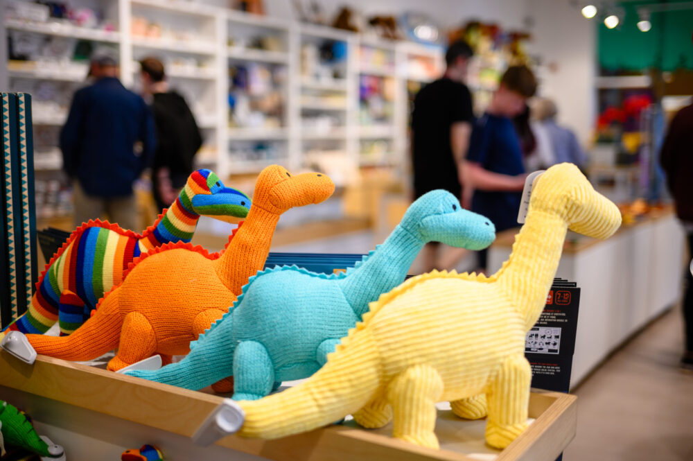 A display of colorful dinosaur plush toys at the World Museum Liverpool during the Dinomania event, with blurred shoppers in the background.